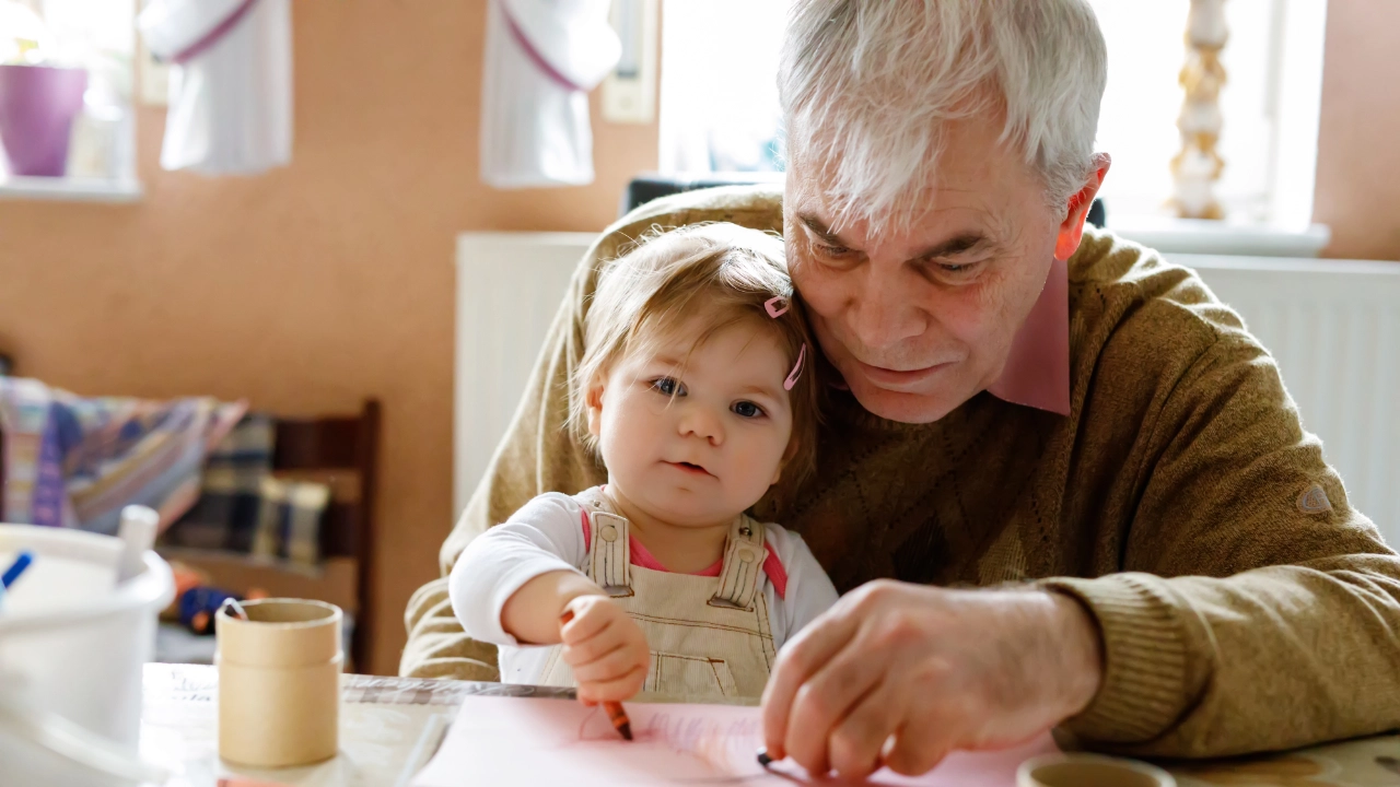 Opa malt mit seiner Enkeltochter ein Bild mit Wachsmalstiften. Sie sitzt dabei auf seinem Schoß.