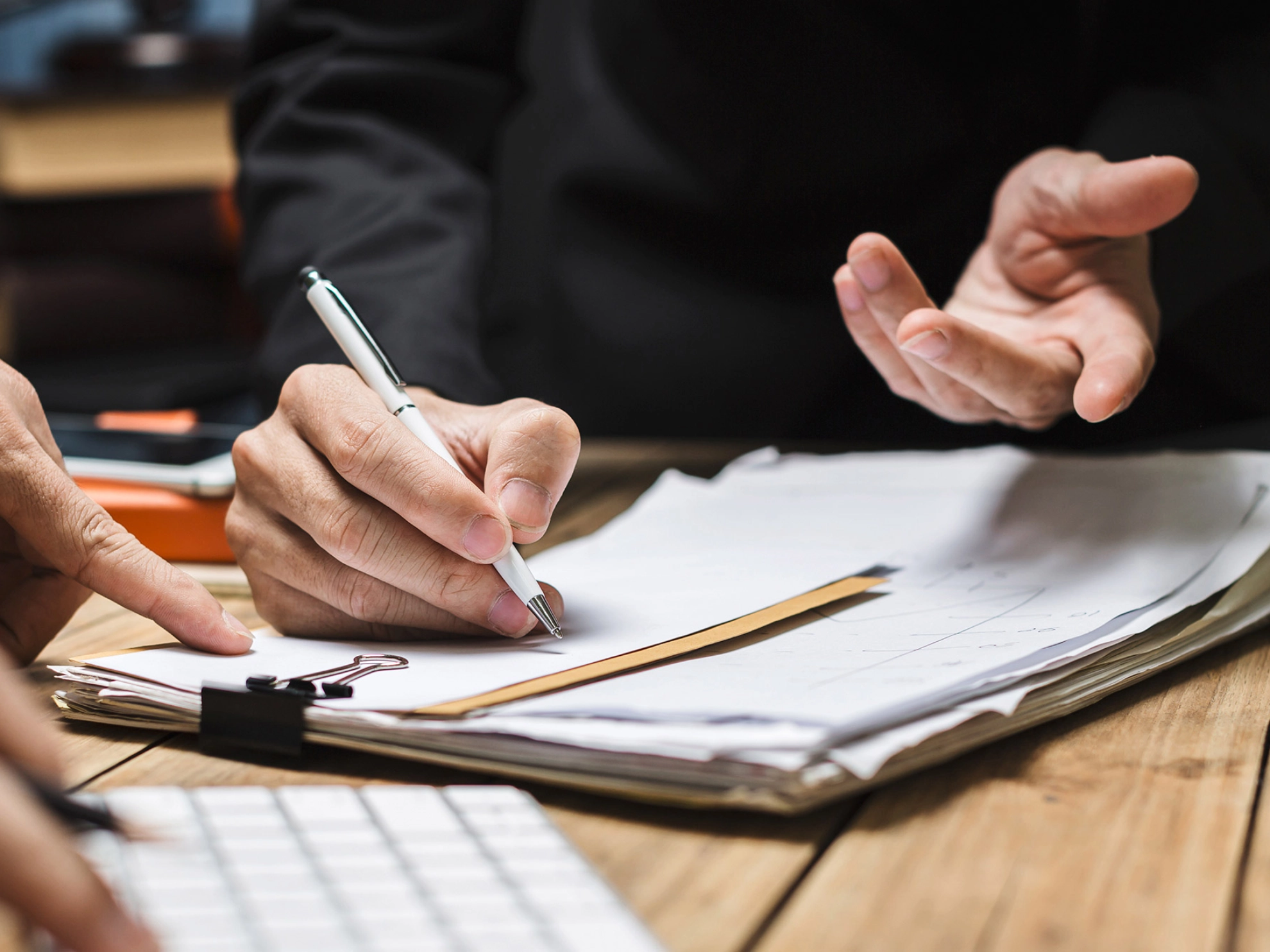 Ein macht Notizen auf Papier auf einem Klemmbrett. Eine andere Person tippt auf einer Tastatur.